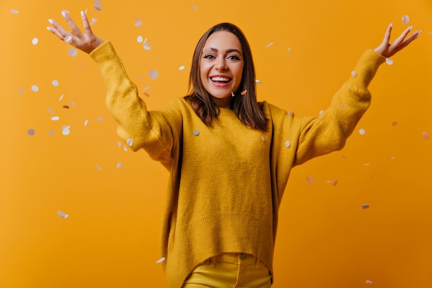 Free photo excited female model in yellow soft sweater standing under confetti. portrait of blithesome woman with straight dark hair.