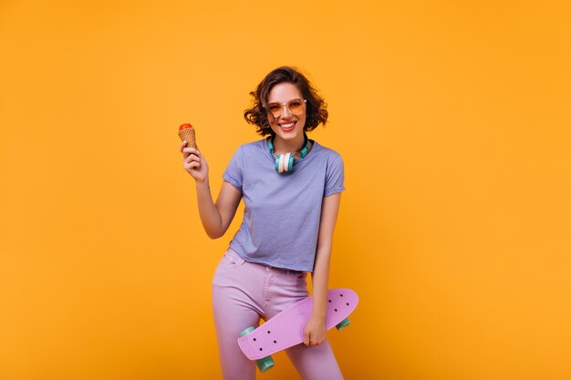 Excited female skateboarder eating ice cream. Indoor photo of ecstatic curly girl with little purple longboard.