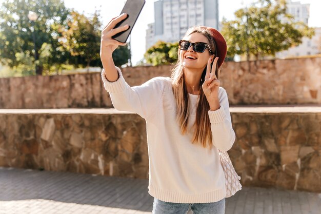 Excited french girl with long-hairstyle making selfie. Outdoor photo of magnificent laughing lady posing with smartphone.