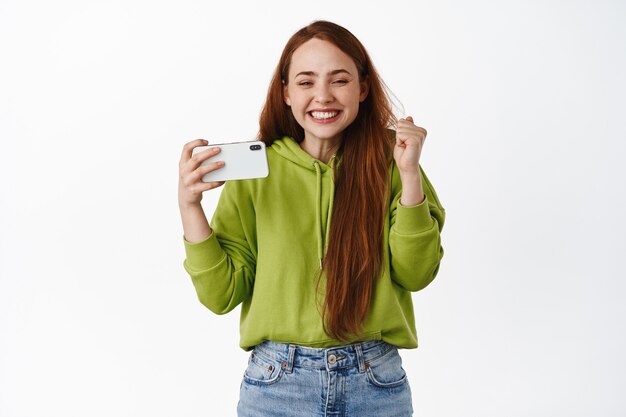 Excited ginger girl holding horizontal smartphone and winning, smiling pleased, triumphing of online achievement, standing on white