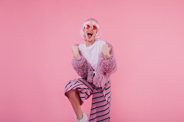 Free Photo excited girl in glamorous pink clothes expressing happiness studio portrait of trendy shorthaired lady wears fluffy jacket and sunglasses