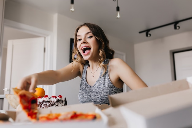 Free photo excited girl holding slice of pizza. blithesome female model enjoying fast food at home.