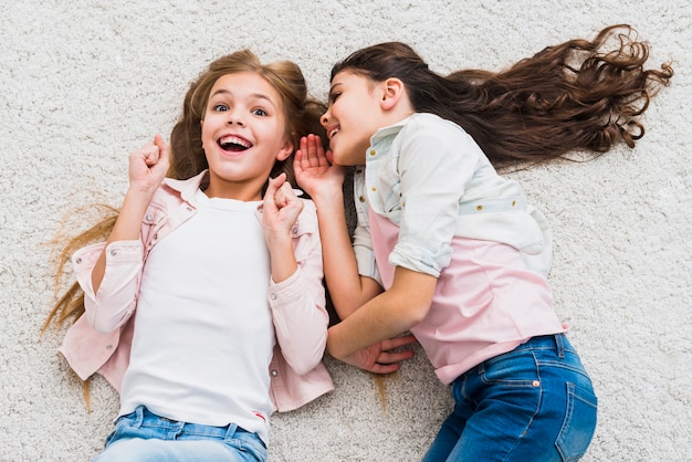 Free photo excited girl listening to friend whispering to her ear lying on carpet