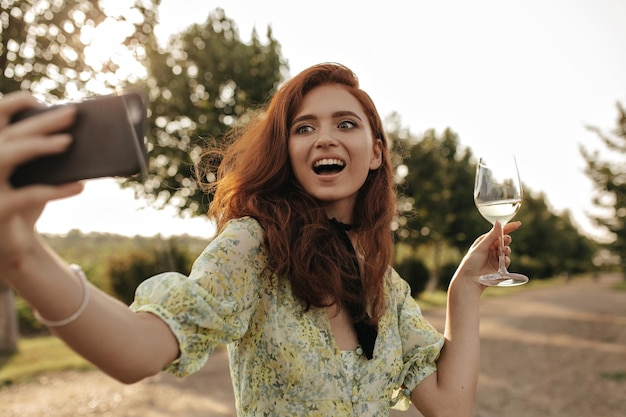 Free Photo excited girl with ginger hair and black bandage on neck in yellow dress holding glass with wine and taking selfie outdoor