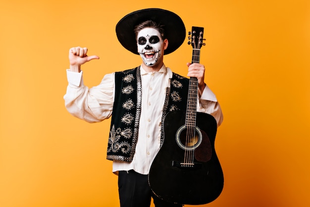 Free photo excited guy in mexican traditional clothes holding guitar. happy dead singer having fun at halloween party.