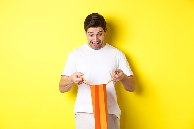 Free Photo excited guy open bag with gift, looking inside with amazement and happy face, standing against yellow background.