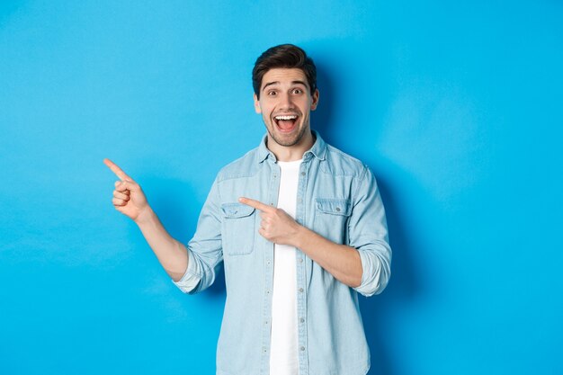 Excited handsome 25s years man with beard, pointing fingers left and smiling amazed, standing against blue background