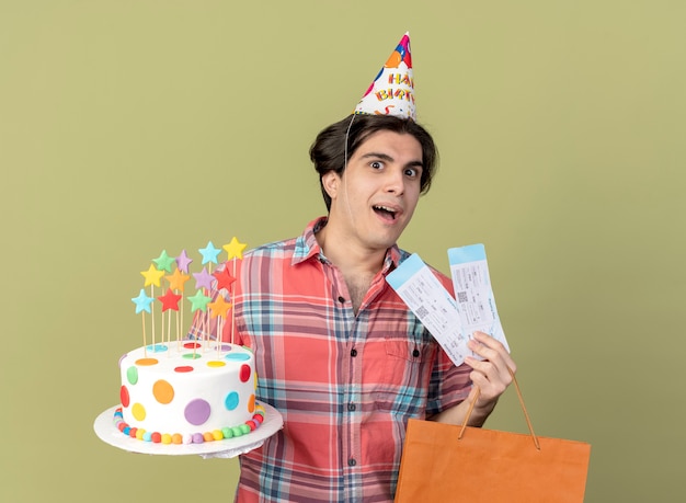 Excited handsome caucasian man wearing birthday cap holds paper shopping bag air tickets and birthday cake 