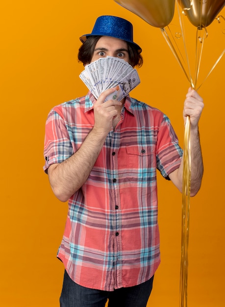 Free photo excited handsome caucasian man wearing blue party hat holds helium balloons and money