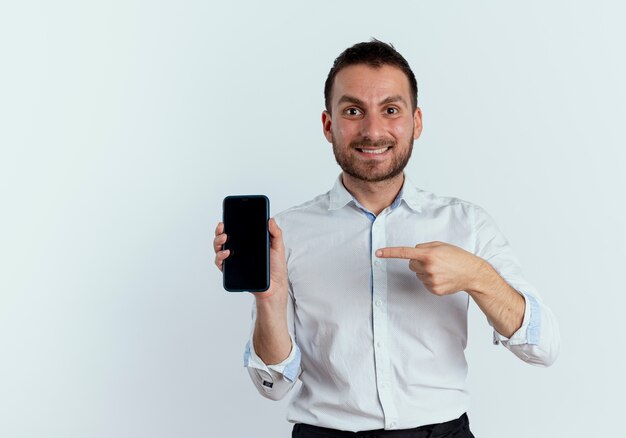 Excited handsome man holds and points at phone isolated on white wall