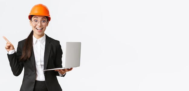 Excited happy asian female engineer industrial woman in safety helmet and business suit showing pres