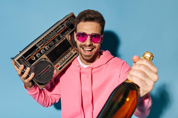 Free photo excited happy bearded man in pink sunglasses and hoodie holds record player and champagne bottle on blue background