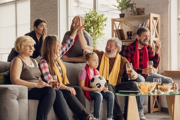 Free photo excited, happy big family watching football, soccer match on the couch at home. fans emotional cheering for favourite national team. having fun from grandparent to children. sport, tv, championship.