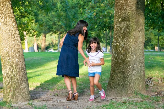 Excited happy girl playing active games with her mom outdoors, standing by trees in park and laughing. Full length. Family outdoor activity and leisure concept