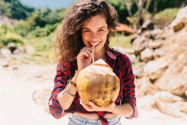 Free Photo excited happy womna dressed shorts and bright shirt sitting on the beach with coconut cocktail