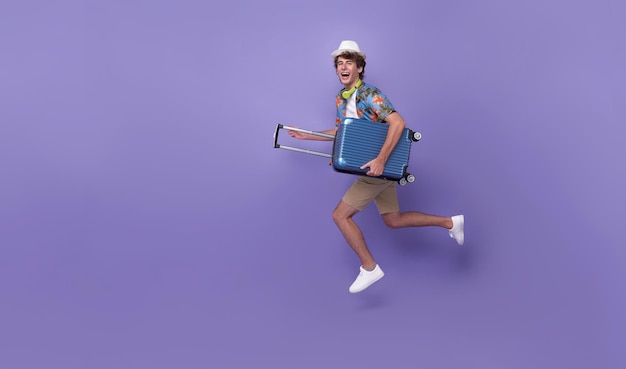 Free photo excited happy young man tourist with luggage jumping isolated on purple studio background