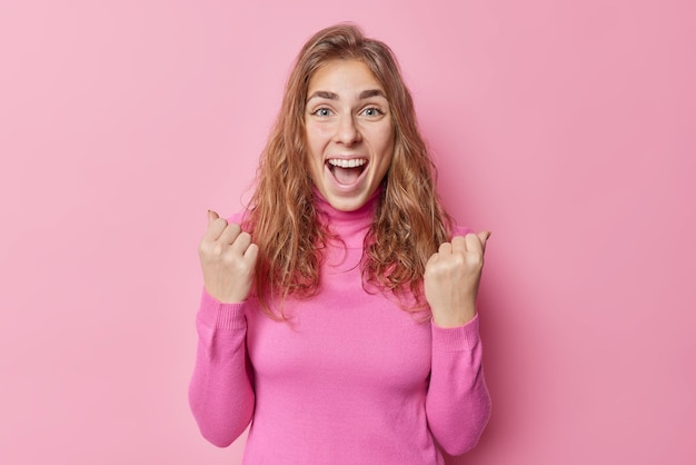 Free photo excited joyful european woman exclaims yes clenches fists triumphs over something celebrates success wears turtleneck isolated over pink background being supportive and happy about something