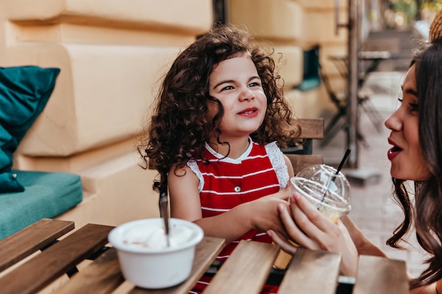 Free photo excited kid have a lunch in cafe. positive little girl sitting in street restaurant with mother.