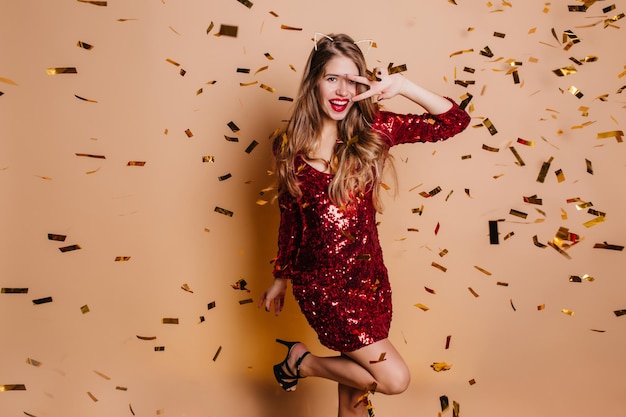 Excited lady with trendy make-up funny posing under sparkle confetti. Wonderful young woman in stylish red dress dancing with smile on beige background at party.