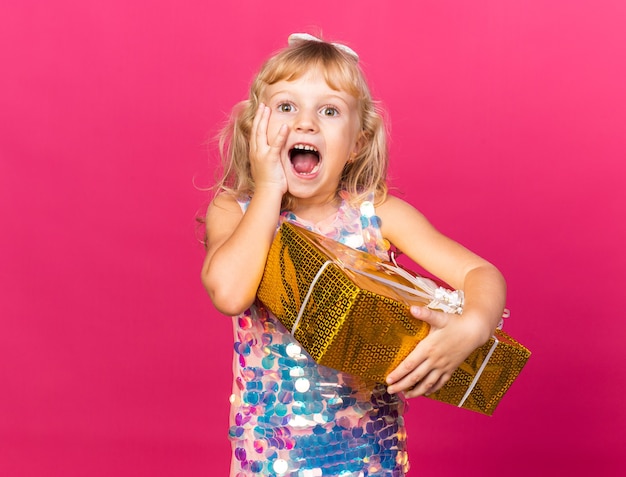 Free photo excited little blonde girl putting hand on face and holding gift box isolated on pink wall with copy space