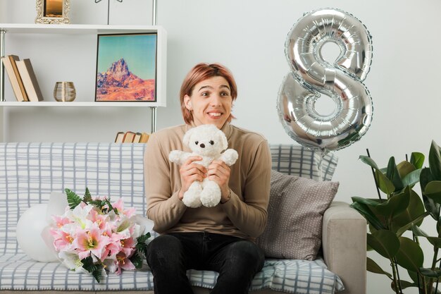 Excited looking side handsome guy on happy women day holding teddy bear sitting on sofa in living room