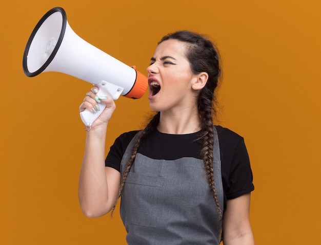 Excited looking at side young female barber in uniform speaks on loudspeaker isolated on orange wall