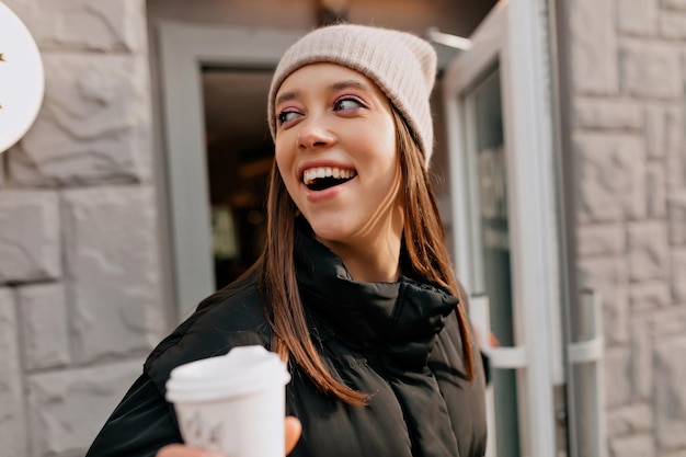 Excited lovely girl with great smile in knitted cap coming out from cafe with coffee to go Pretty charming lady walking around the city in sunny warm spring day