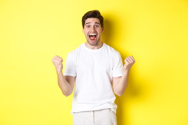 Excited and lucky man winning, clench fists and looking happy, triumphing and celebrating, standing over yellow background.