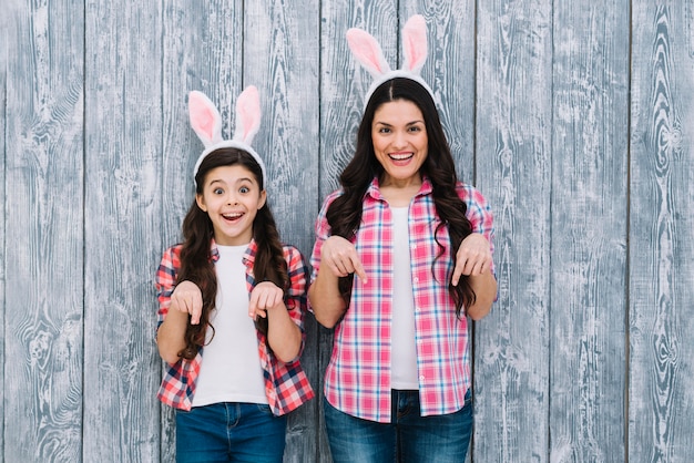 Free photo excited mother and daughter with bunny ears pointing the finger downward against wooden backdrop