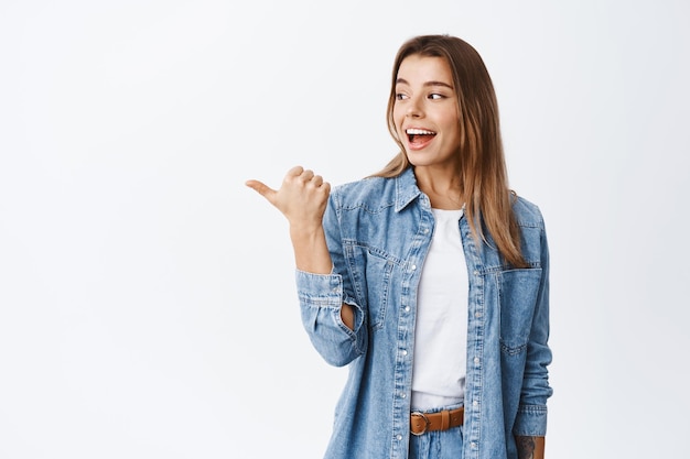 Excited pretty millennial girl turn head and pointing thumb left, checking out good promo offer, demonstrating advertisement on copy space, white