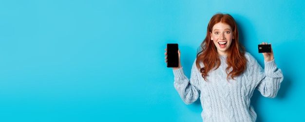 Free Photo excited redhead girl showing mobile phone screen and credit card demonstrating online store or appli