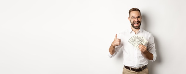 Free photo excited rich man holding money showing thumb up in approval standing over white background