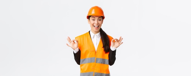 Excited and satisfied asian female chief engineer pleased with amazing work showing okay gesture assure clients guarantee perfect quality standing in safety helmet over white background