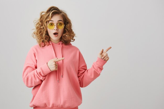 Excited stylish curly-haired girl in sunglasses pointing right, showing way