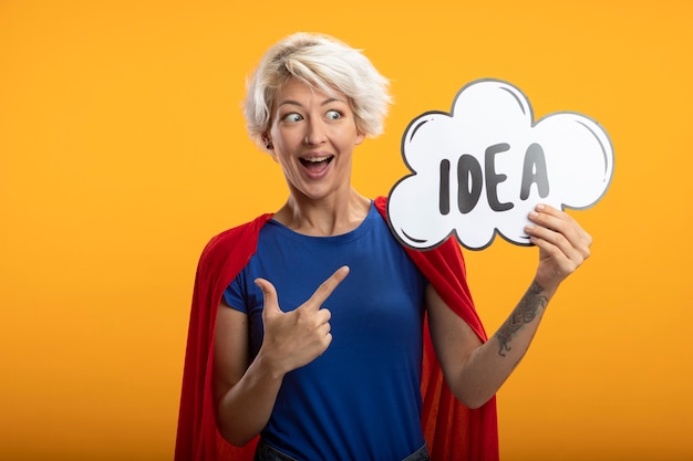 Free photo excited superwoman with red cape holds and points at idea bubble isolated on orange wall