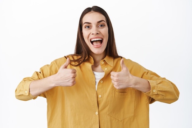 Excited and supportive girlfriend shows thumbs up, stay positive, rooting, you can do it gesture, keep up the good work, well done sign, standing satisfied against white wall