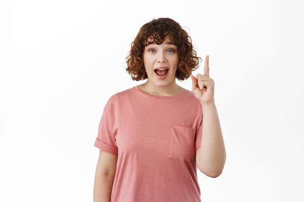 Excited and surprised girl scream from amazement, pointing finger up, say wow, checking out advertisement on top, standing in t-shirt against white background