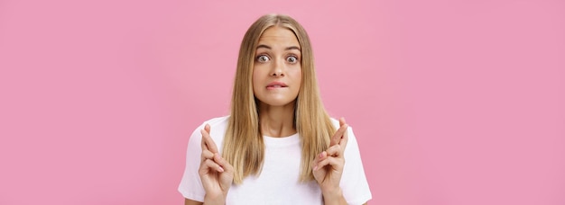 Free photo excited and thrilled attractive emotive fairhaired female with pimples on forehead biting lower lips crossing fingers for good luck making wish praying desire become reality over pink wall