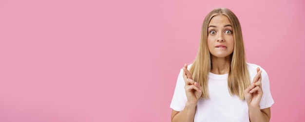 Free photo excited and thrilled attractive emotive fairhaired female with pimples on forehead biting lower lips crossing fingers for good luck making wish praying desire become reality over pink wall