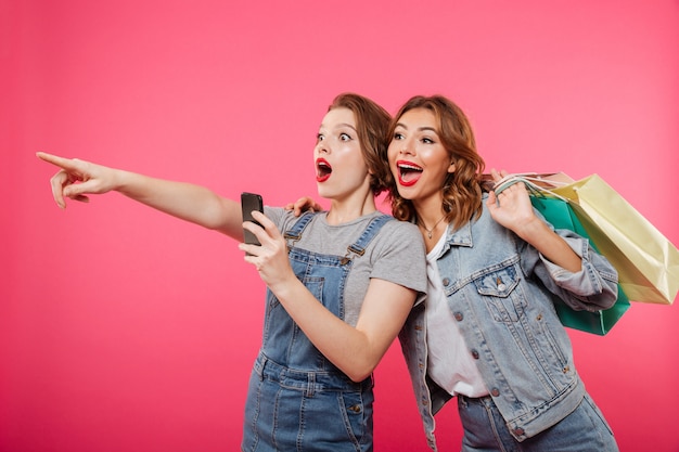 Excited two women friends holding shopping bags using mobile phone.