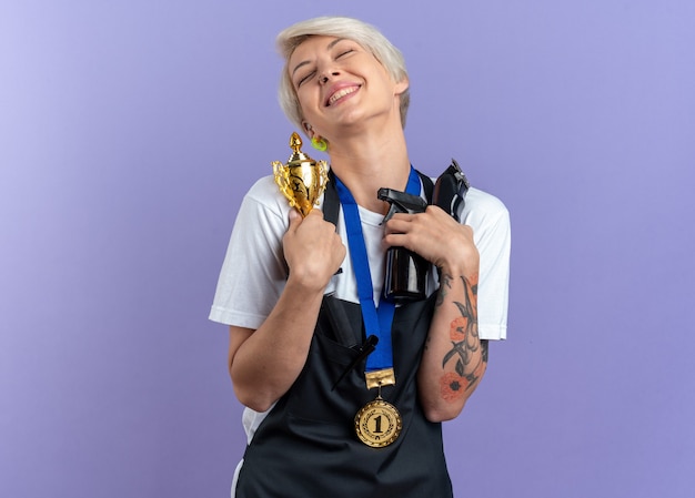 Free photo excited with closed eyes tilting head young beautiful female barber in uniform wearing medal holding barber tools and winner cup isolated on blue wall