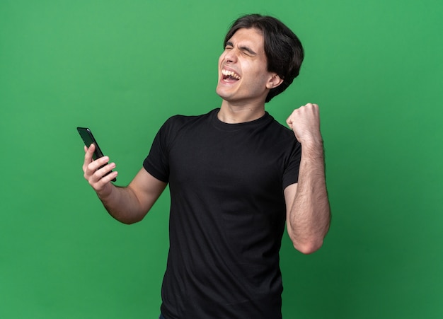 Excited with closed eyes young handsome guy wearing black t-shirt holding phone showing yes gesture isolated on green wall