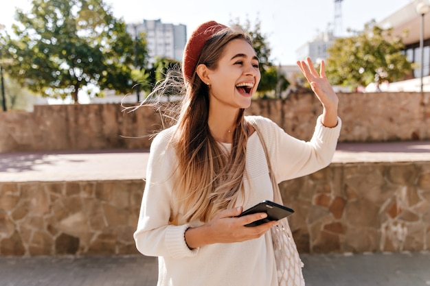 Excited woman in cardigan posing on the street. Long-haired appealing girl in red beret waving her hand.