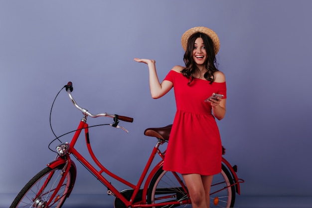 Free photo excited woman with phone in hand standing beside her bike . emotional brunette girl in straw hat posing in front of bicycle.