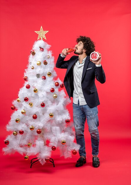 Excited young adult holding clock and and drinking a glass of wine and standing near Xsmas tree on red stock photo