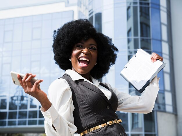 Excited young african businesswoman holding mobile phone and clipboard