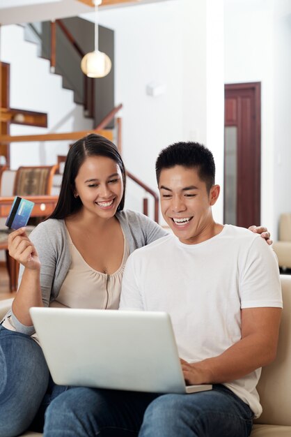 Excited young Asian couple sitting on couch at home with laptop and credit card