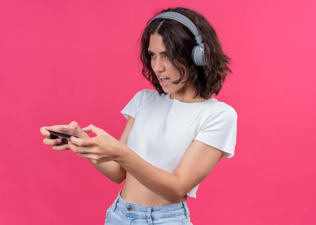 Excited young beautiful woman wearing headphones and holding mobile phone looking at left side on pink wall