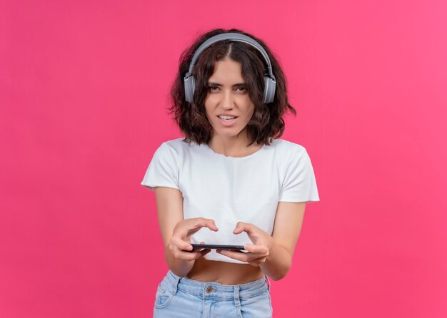 Excited young beautiful woman wearing headphones and holding mobile phone on pink wall with copy space