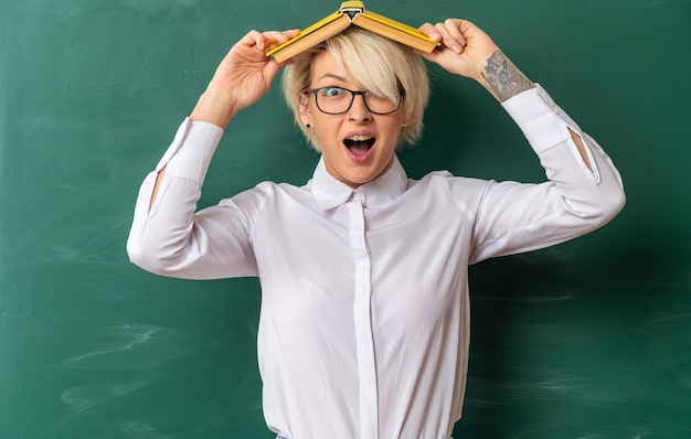 Free photo excited young blonde female teacher wearing glasses in classroom standing in front of chalkboard holding book on head looking at camera